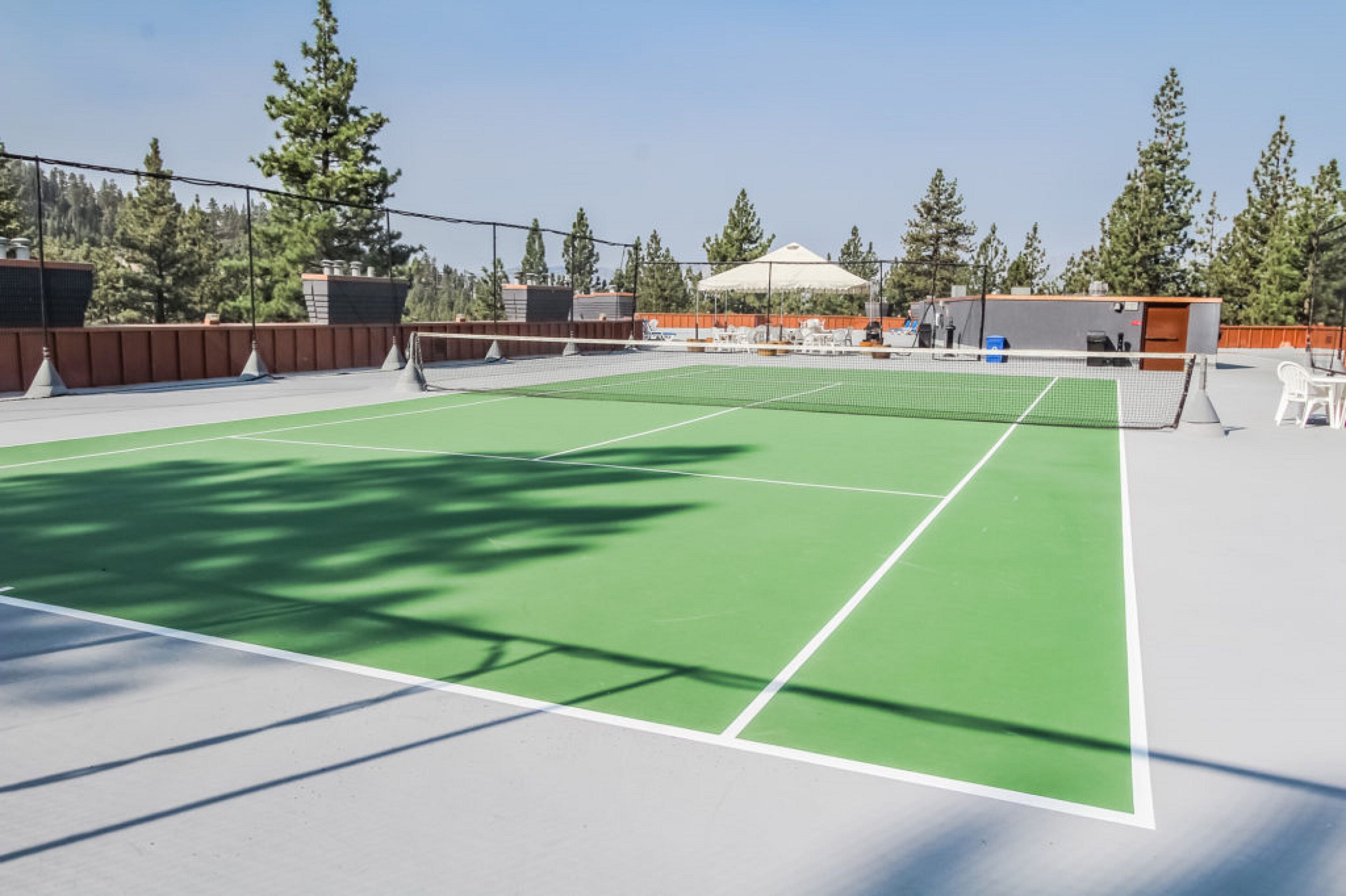 Una cancha de tenis exterior con superficie verde, rodeada de árboles y una zona de estar techada.