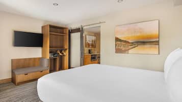 Una habitación de hotel con una cama grande, un banco de madera, un televisor montado en la pared, una zona de cocina y un cuadro de un atardecer sobre un puente.
