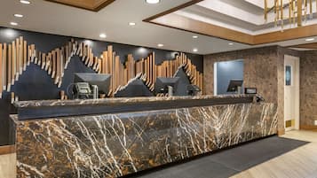 A hotel reception desk with a marble counter, a modern wooden wall design, and a staircase in the background.