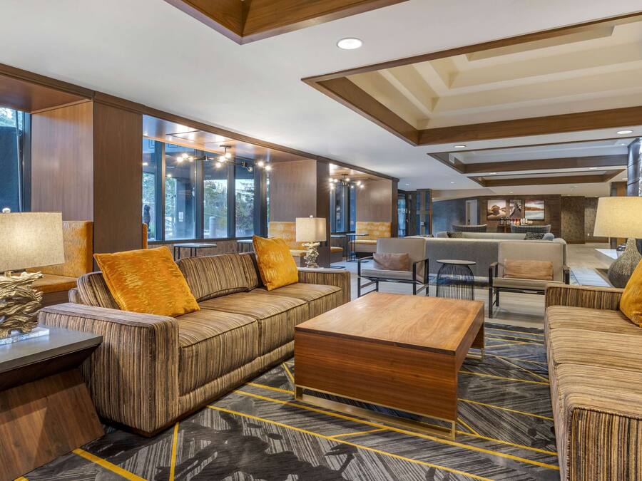 Un moderno hall de hotel con un área de estar central que presenta sofás a rayas y una mesa de centro de madera. Hay varias lámparas y un adorno sobre una mesita auxiliar. La habitación tiene ventanas grandes y vistas al exterior.