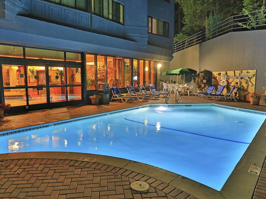 A swimming pool with lounge chairs and umbrellas, surrounded by a building with large windows and a brick pathway.