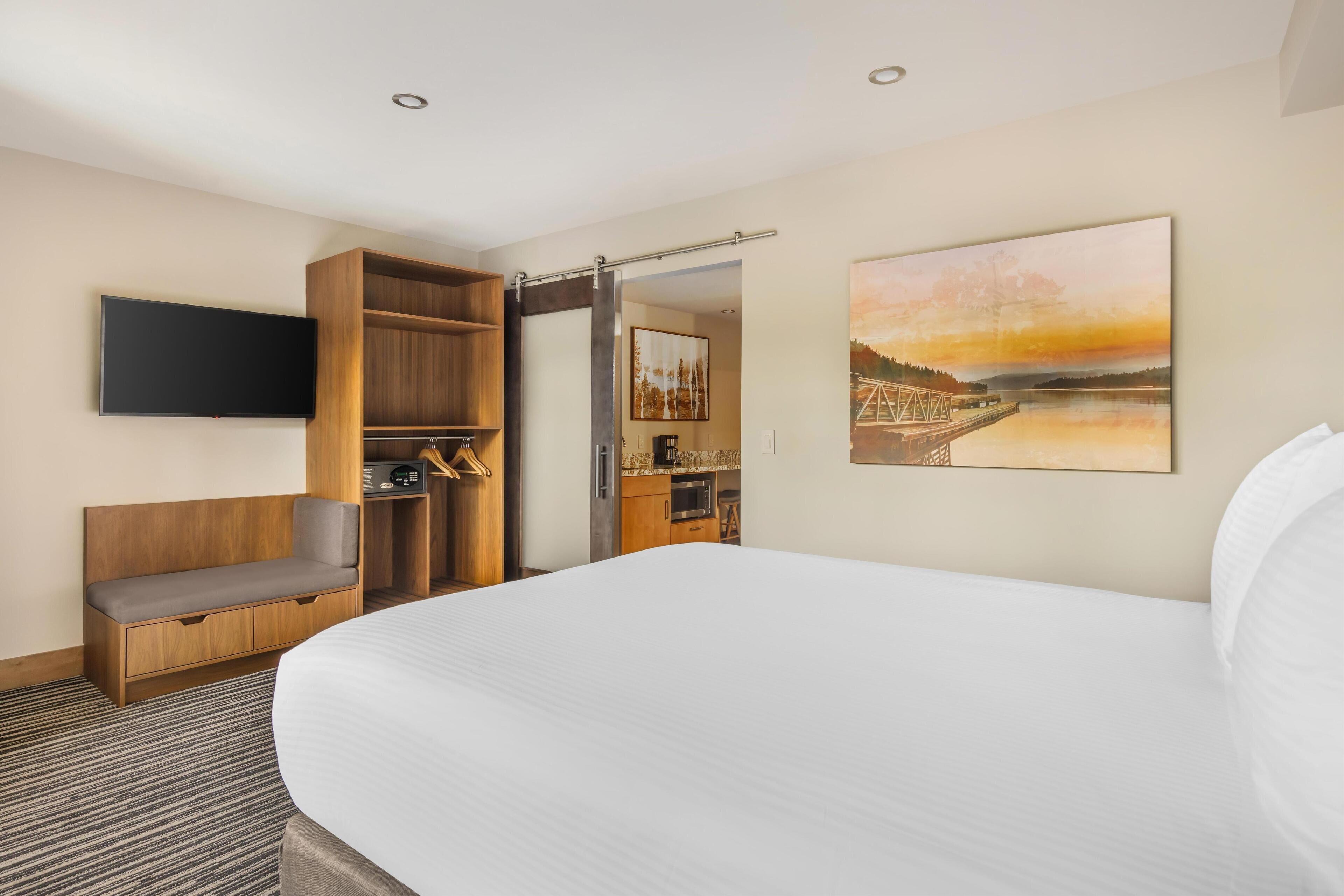 A hotel room with a large bed, a wooden bench, a TV mounted on the wall, a kitchen area, and a painting of a sunset over a bridge.