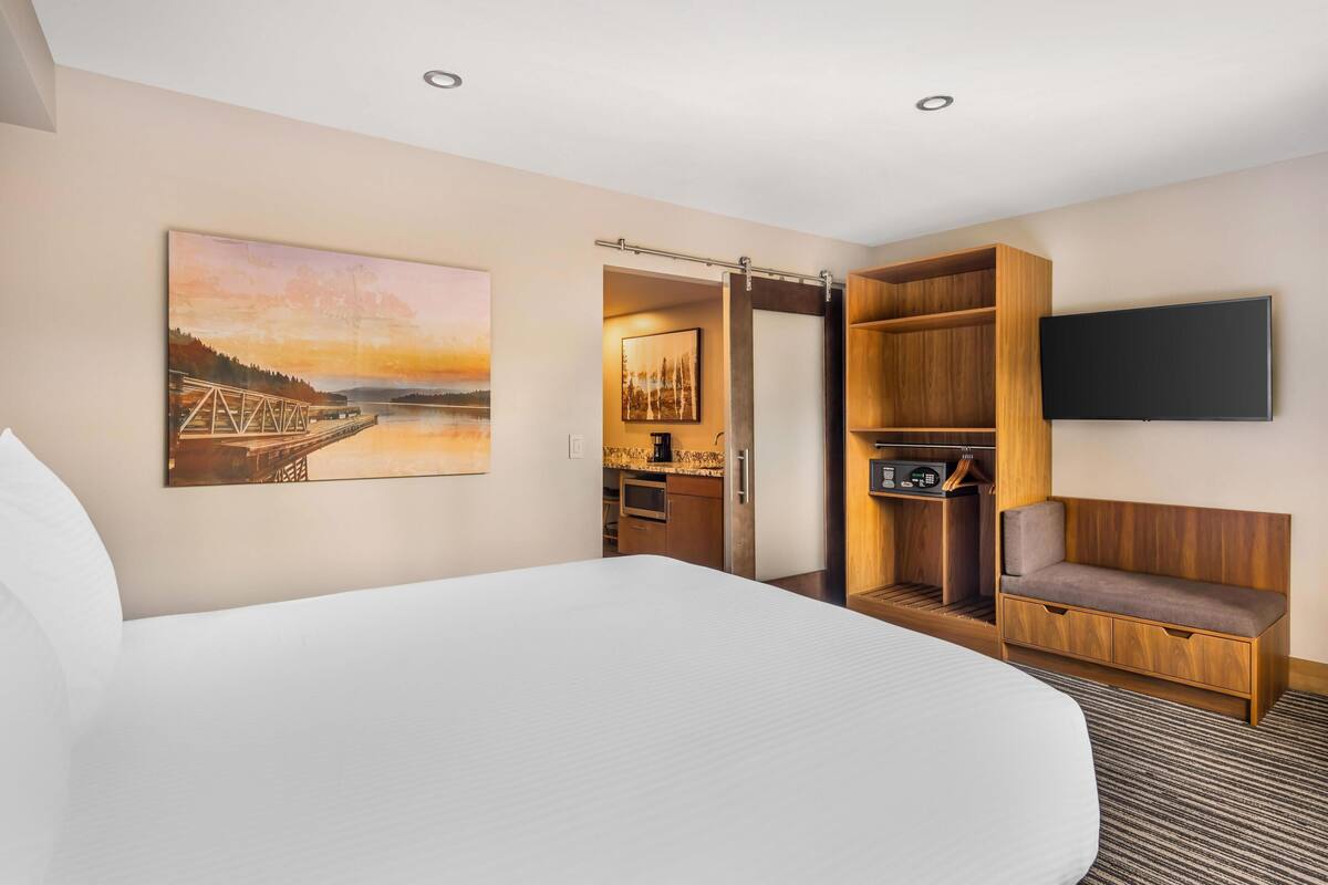 Habitación de hotel con una cama grande, un cuadro de un puente, un televisor montado en la pared, un banco de madera y una pequeña cocina.