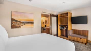 Habitación de hotel con una cama grande, un cuadro de un puente, un televisor montado en la pared, un banco de madera y una pequeña cocina.