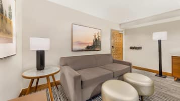 A modern hotel room with a grey sofa, two ottomans, a small round table with a lamp, a large framed picture on the wall, and a wooden cabinet.