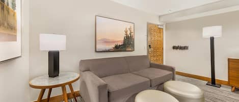 Una habitación de hotel moderna con un sofá gris, dos otomanas, una pequeña mesa redonda con una lámpara, un cuadro grande con marco y un armario de madera.