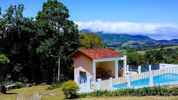 A small, single-story building with a red-tiled roof, a swimming pool, and a view of rolling hills and clouds in the background. - Chácara do Alemão - Recanto Jd.Silvestre (Vargem)