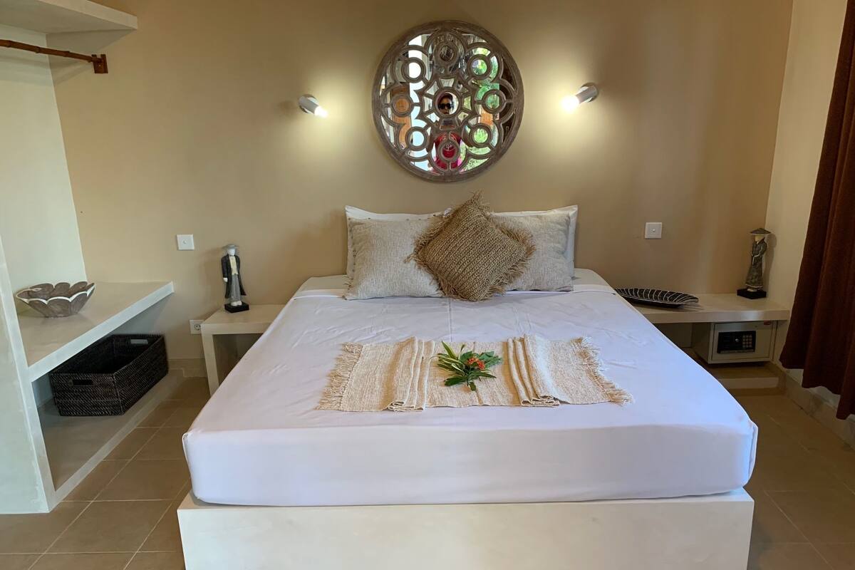 A neatly made bed with a decorative headboard, a wall-mounted light fixture, and a small statue on the nightstand.