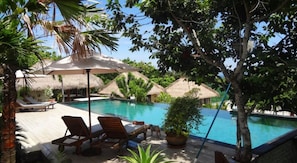 A pool area with lounge chairs and umbrellas, surrounded by thatched-roof huts and lush greenery. - Villa Anjing (Nusa Dua)