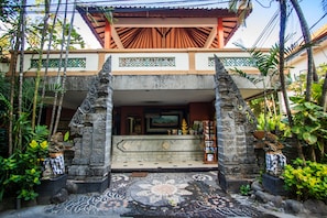 A traditional building with a tiled entrance, a stone balustrade, and a tiled floor leading to a covered area with a television. - Sri Ratu Hotel (Legian)