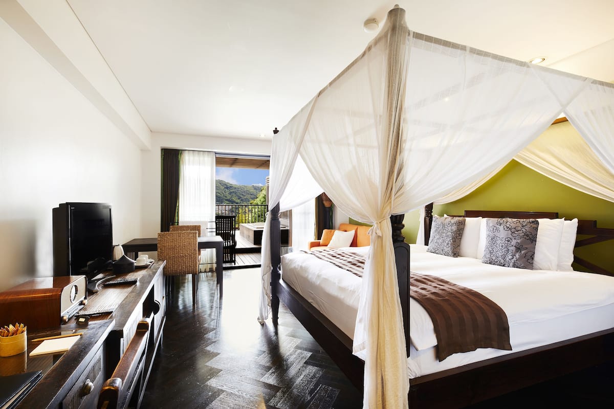 A four-poster bed with a canopy, a desk with a computer, and a view of a green landscape through the window.