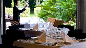 Une table de restaurant dressée avec une nappe blanche, des couverts en argent et des verres à vin, avec un serveur en arrière-plan.