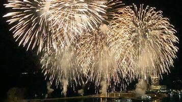 Paisatge urbà nocturn amb un espectacle de focs artificials sobre l'aigua, edificis al primer pla i un pont al fons.