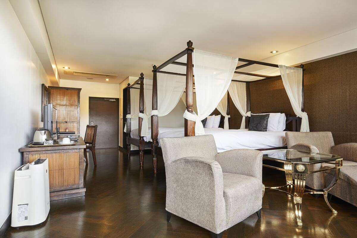 A spacious hotel room with a four-poster bed, a sitting area with two armchairs and a small table, a desk with a chair, and a large mirror.