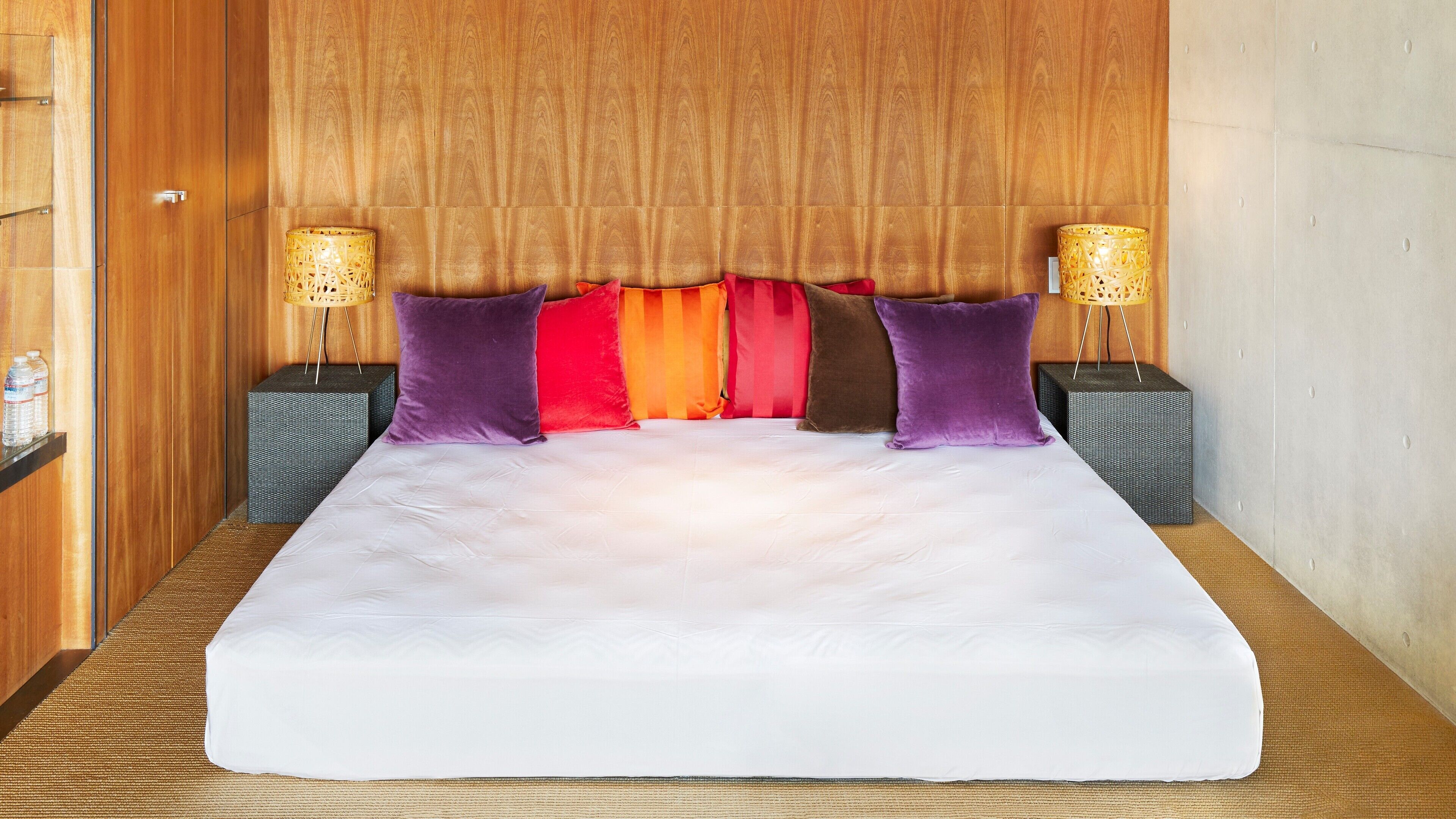 A large bed with a white mattress, surrounded by purple and red pillows, and two nightstands with lamps.