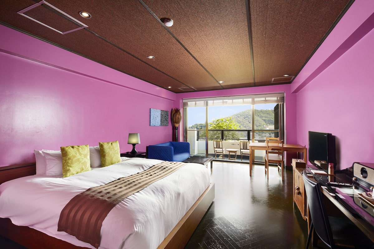 A modern hotel room with a large bed, a desk, a TV, and a balcony with a view of the mountains.