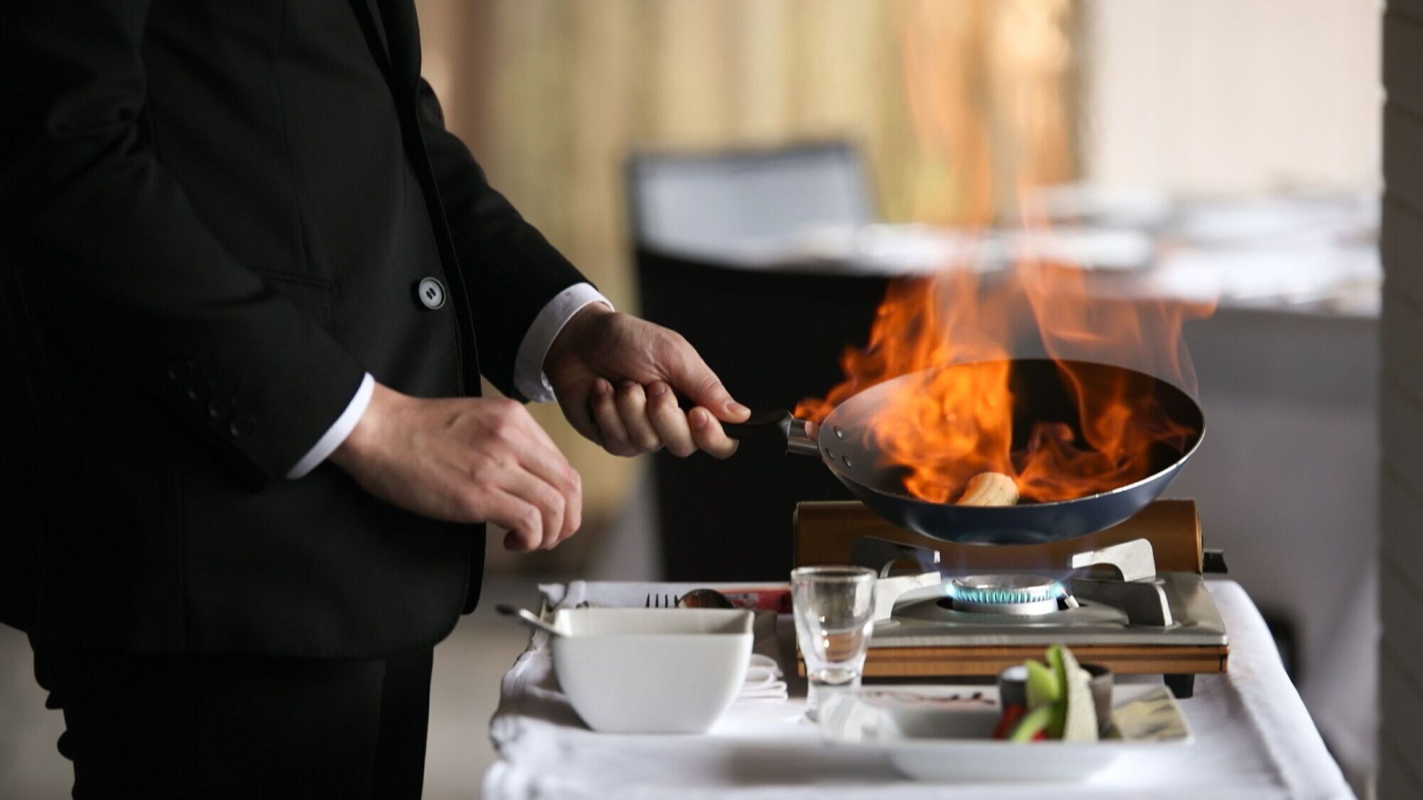 A person in a black suit cooking with a wok on a portable stove.