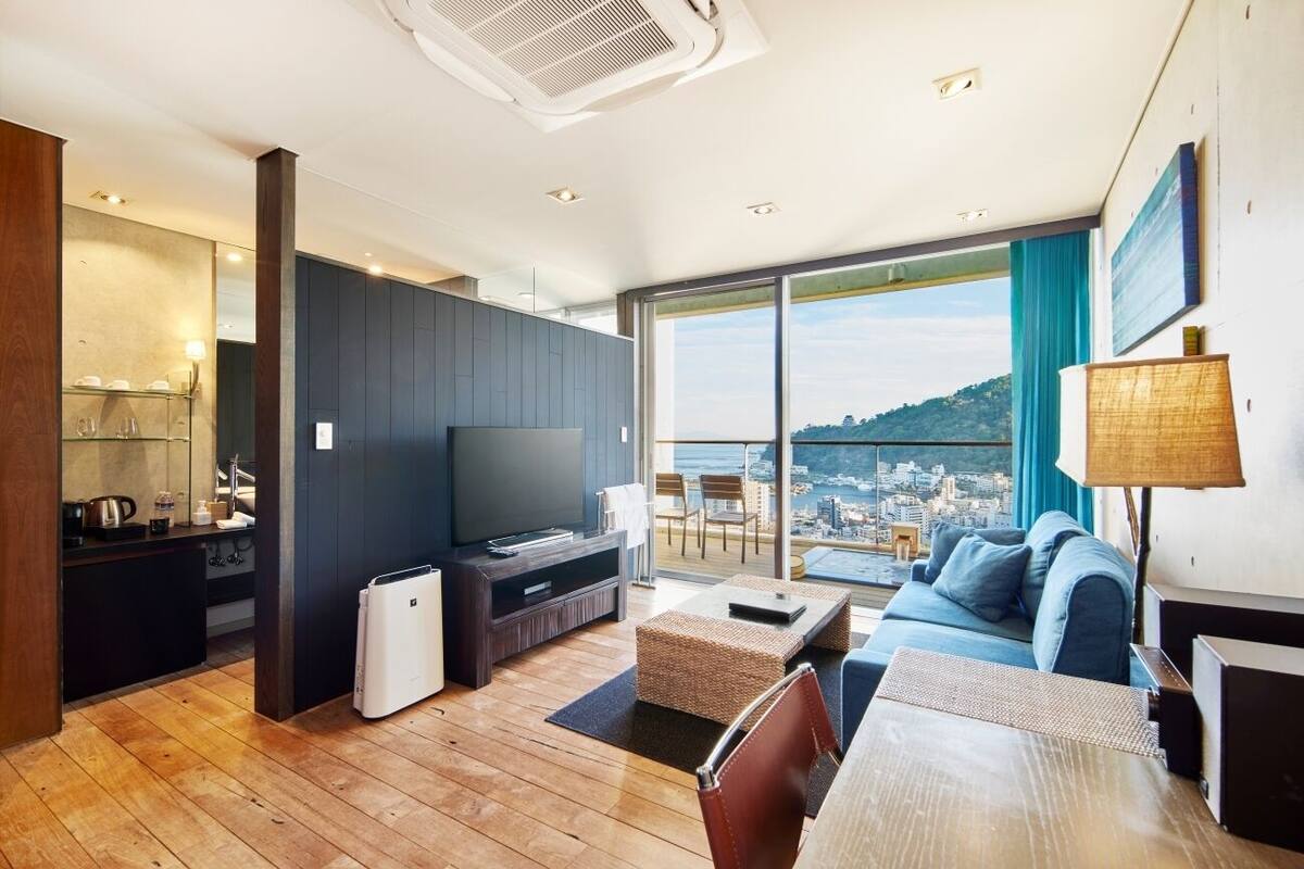A modern living room with a large TV, a sofa, a dining table, and a balcony with a city view.