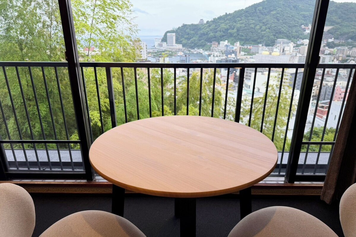 A round table with two chairs in a room with a view of a cityscape and greenery through large windows.