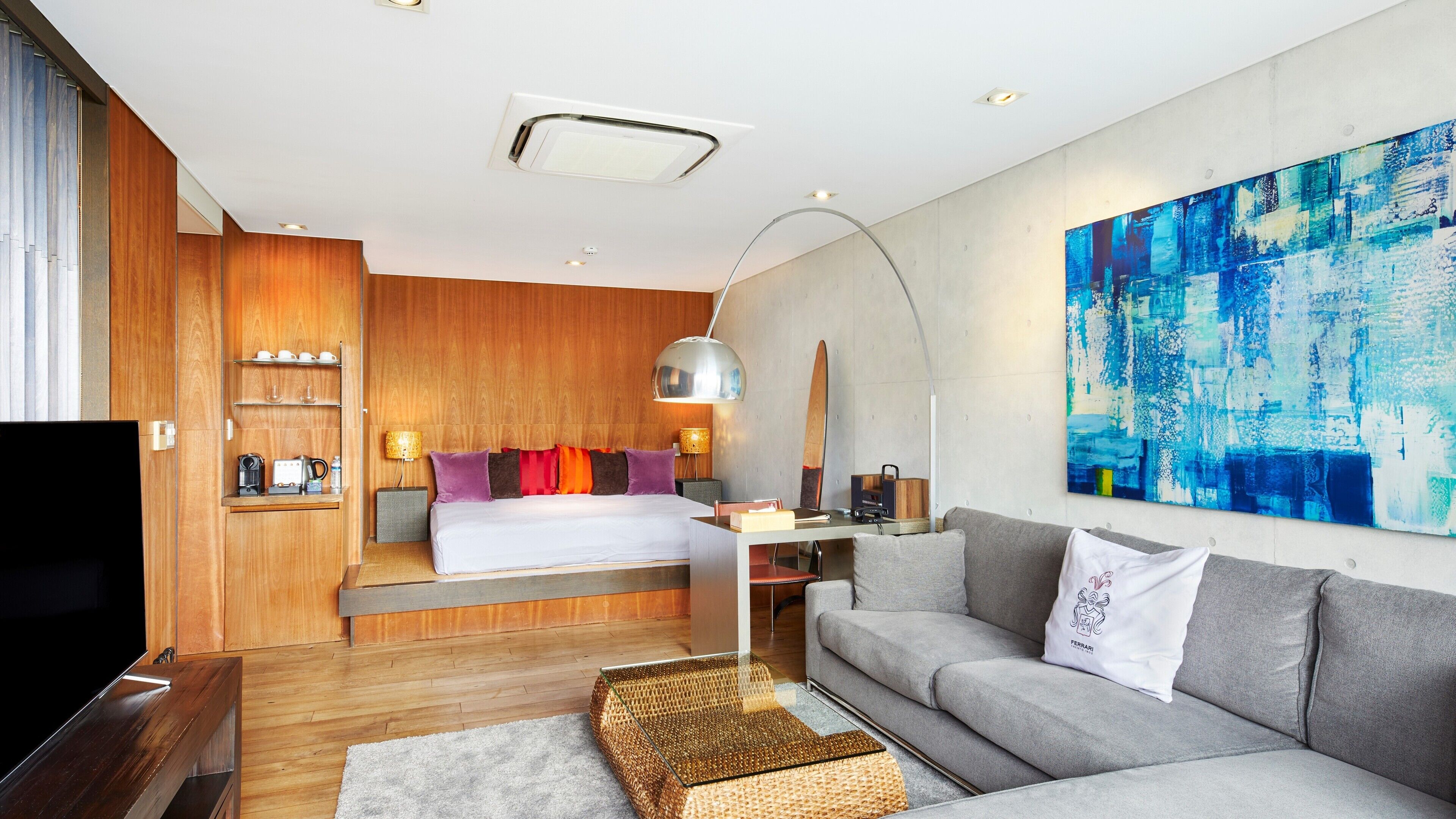 Ein modernes Hotelzimmer mit einer Wand in Holzoptik, einem großen Bett mit bunten Kissen, einem grauen Sofa, einem Couchtisch und einem großen abstrakten Gemälde.