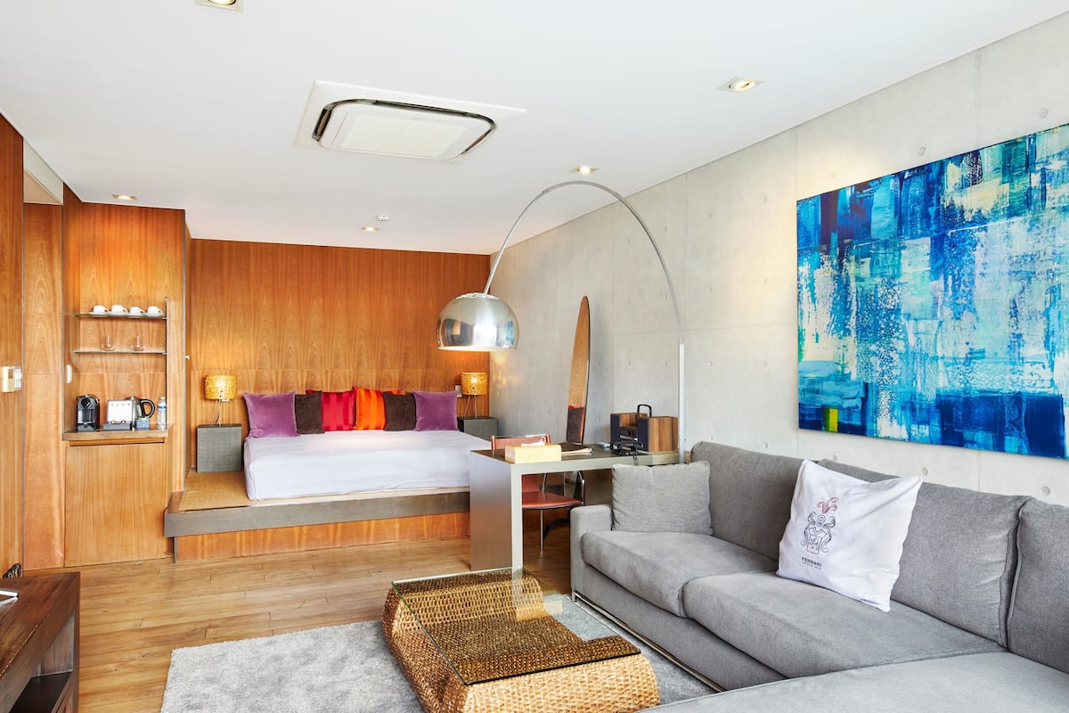 A modern hotel room with a wooden accent wall, a large bed with colorful pillows, a grey sofa, a coffee table, and a large abstract painting on the wall.