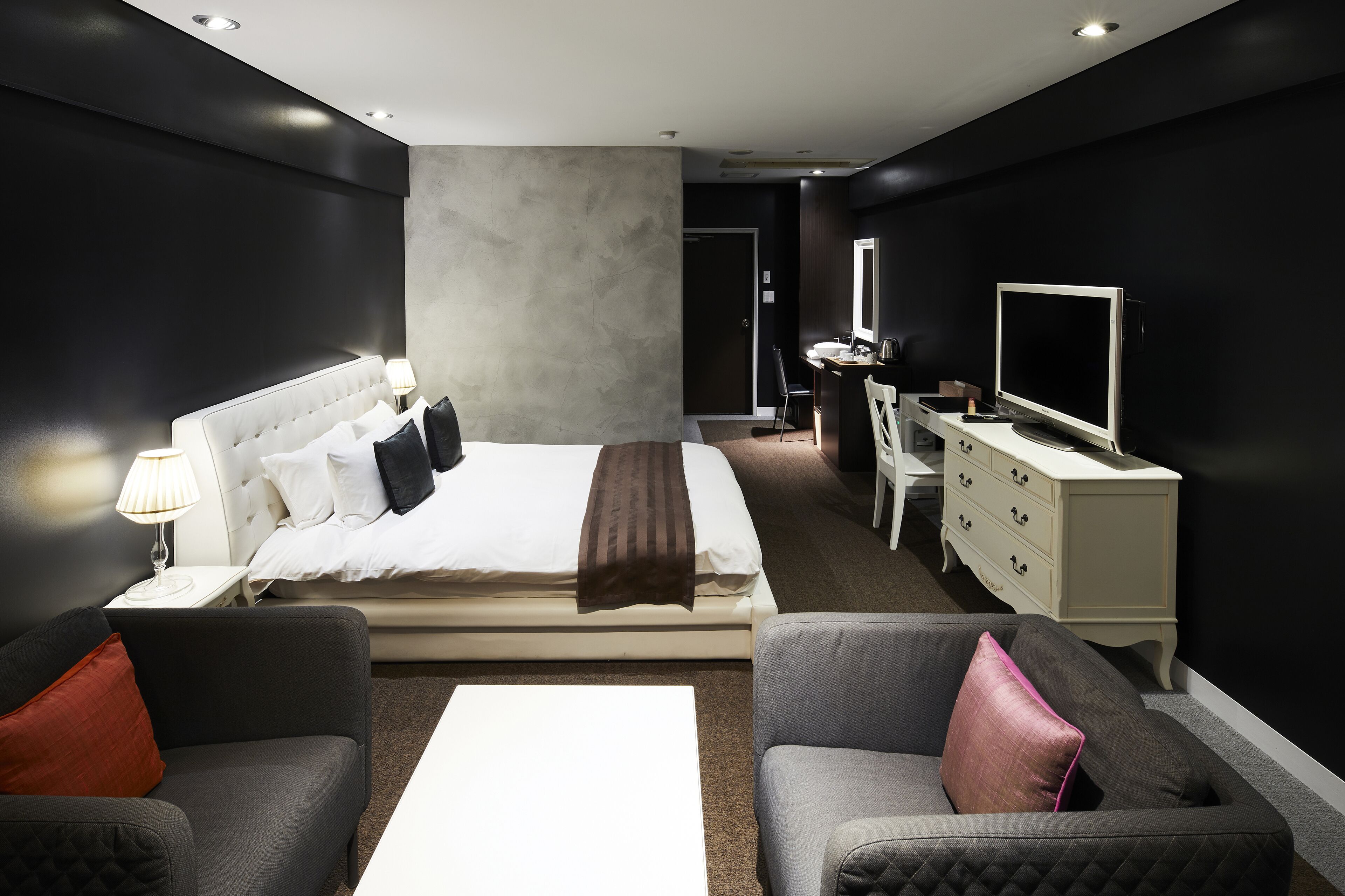 A modern hotel room with a large bed, a desk, a TV, and two armchairs.