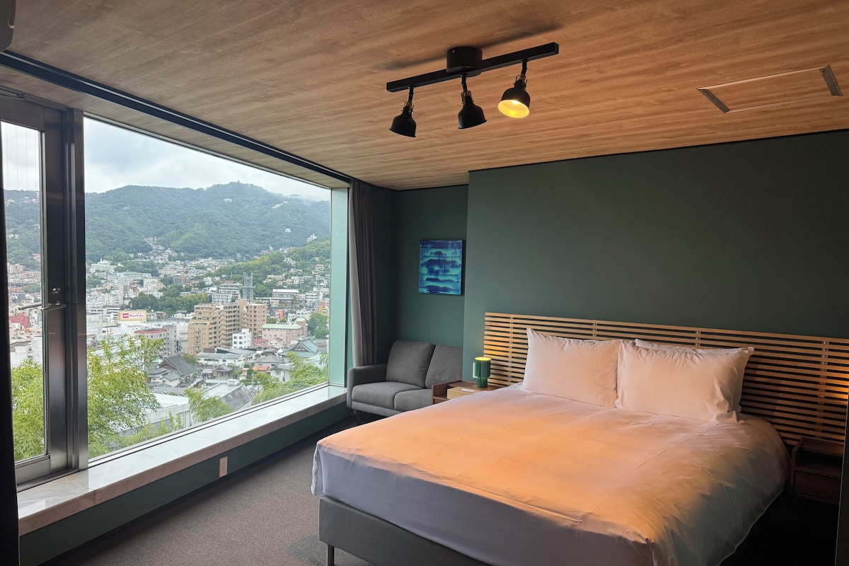 Une chambre d’hôtel avec une grande fenêtre, un lit, une chaise et une vue sur le paysage urbain.