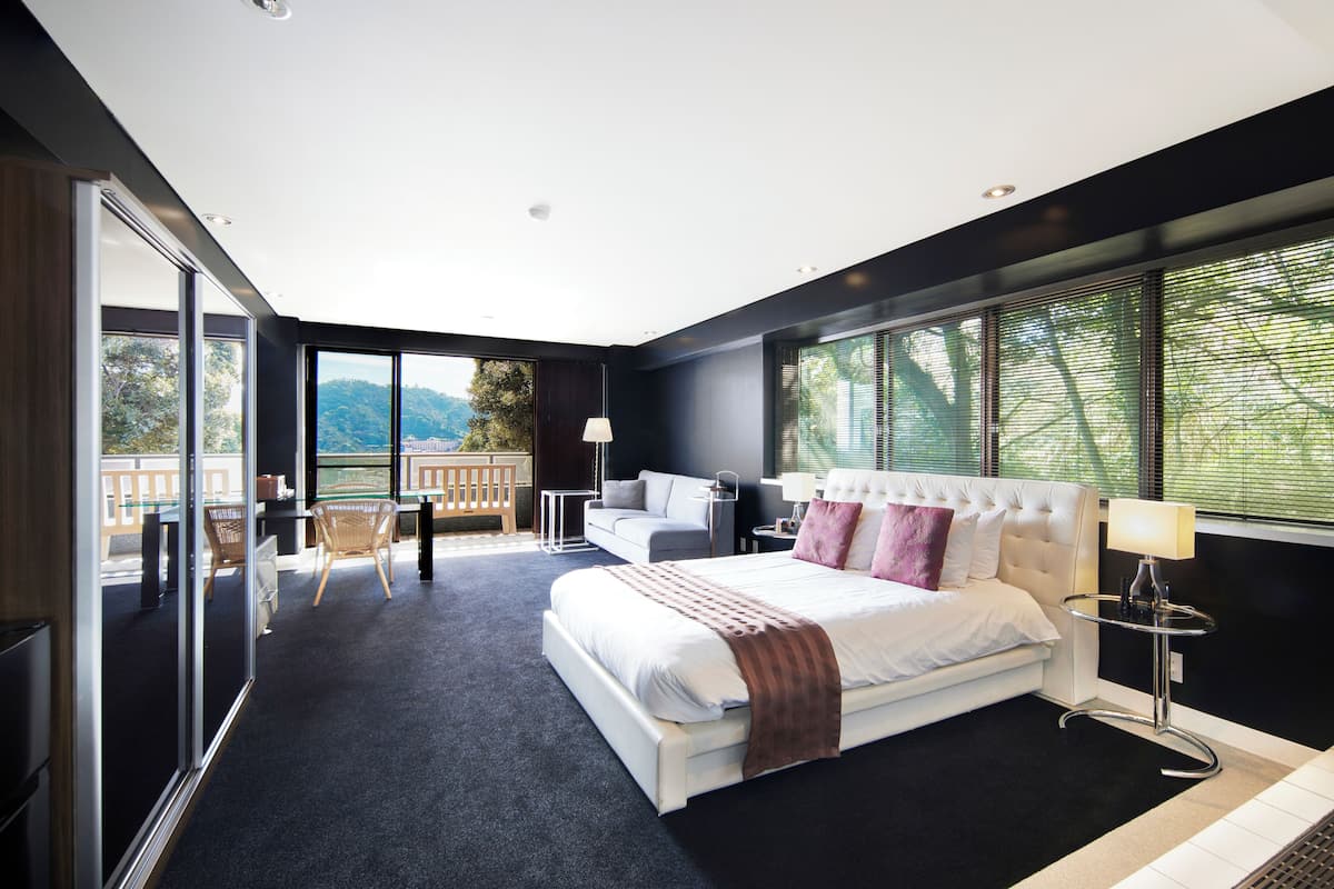 A modern hotel room with a large bed, a sitting area with a sofa and chairs, a small table, and a balcony with a view of the outdoors.