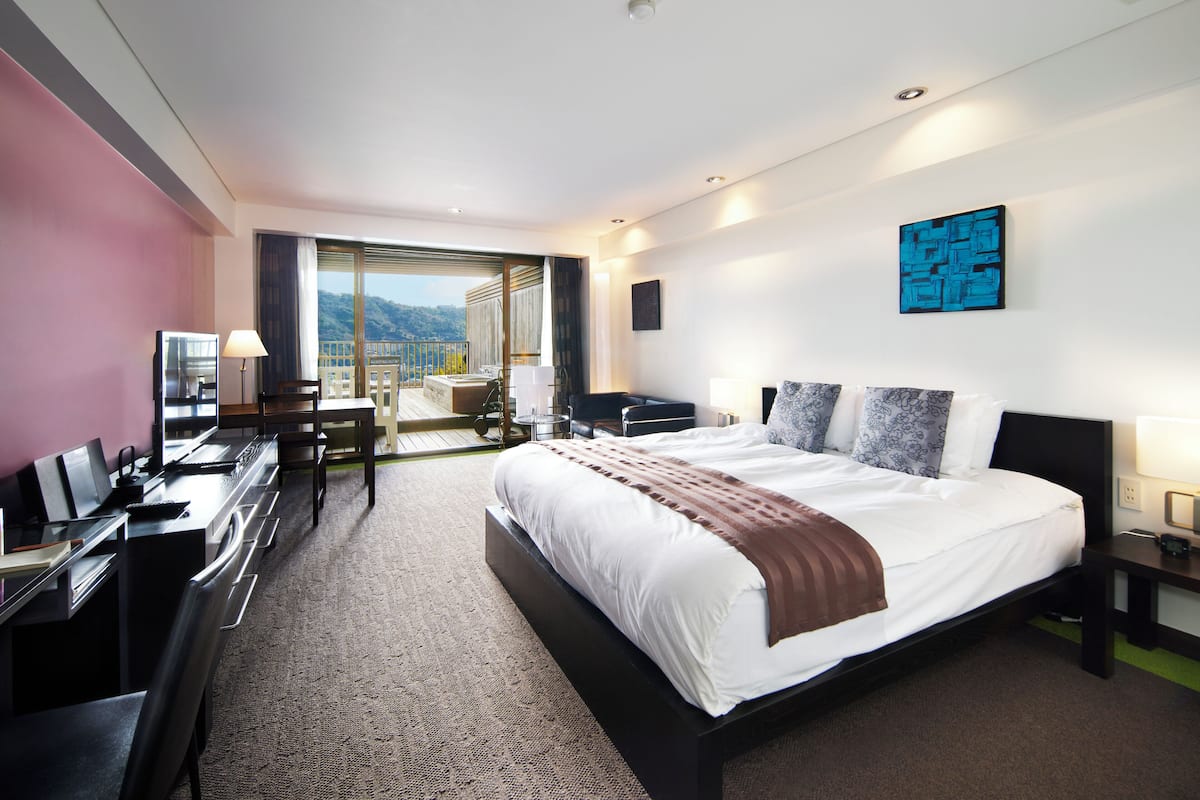 A modern hotel room with a large bed, a desk, and a chair. There is a balcony with a view of the mountains.