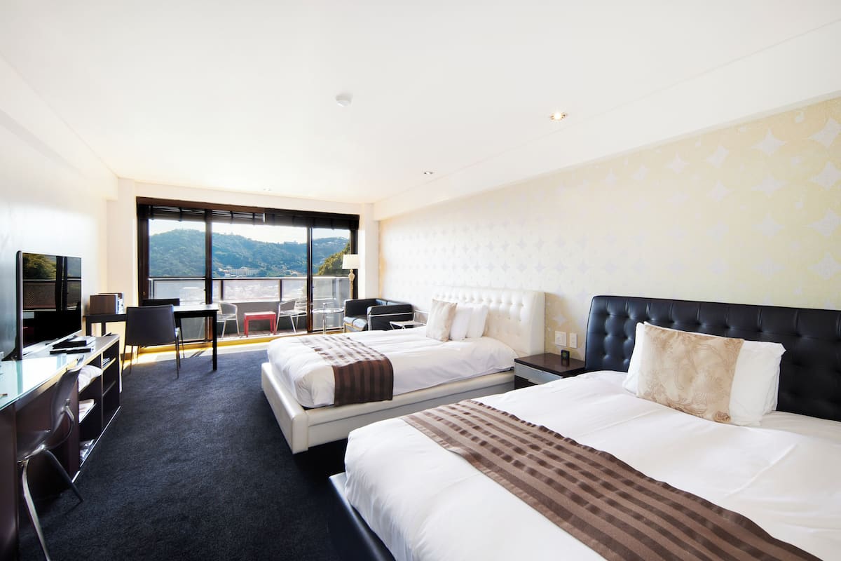 A hotel room with two beds, a desk, a chair, and a large window offering a view of a mountainous landscape.