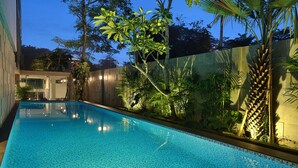 A rectangular swimming pool with clear blue water, surrounded by lush greenery and a modern building facade.