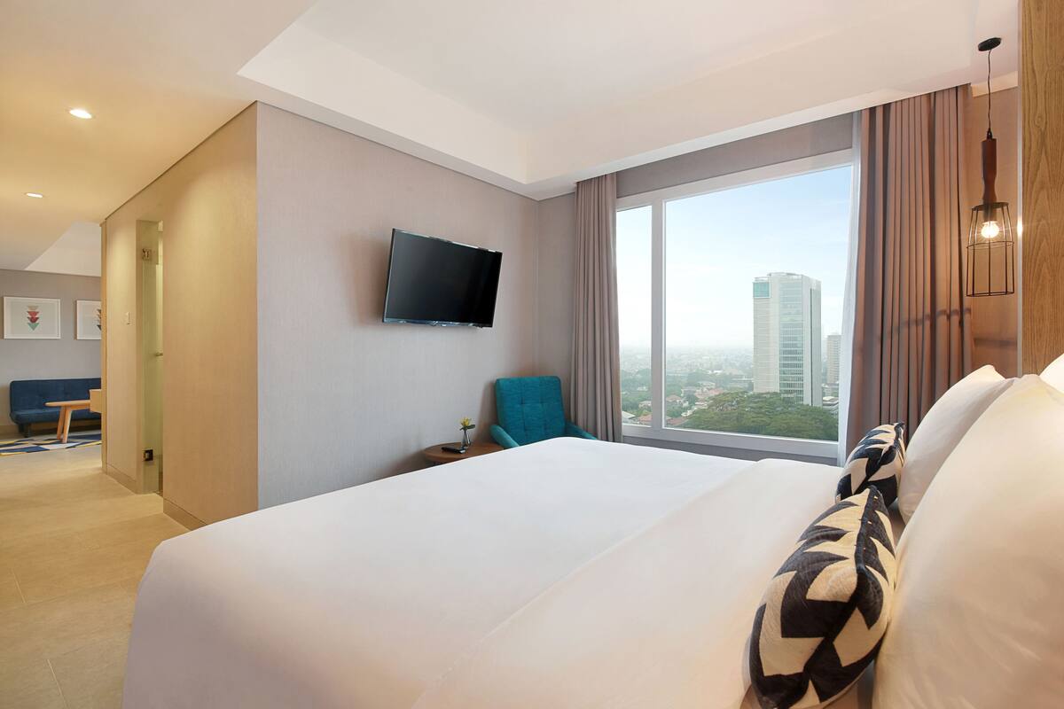 Une chambre d’hôtel avec un grand lit, un téléviseur fixé au mur, une vue sur la ville par une grande fenêtre et un fauteuil bleu.