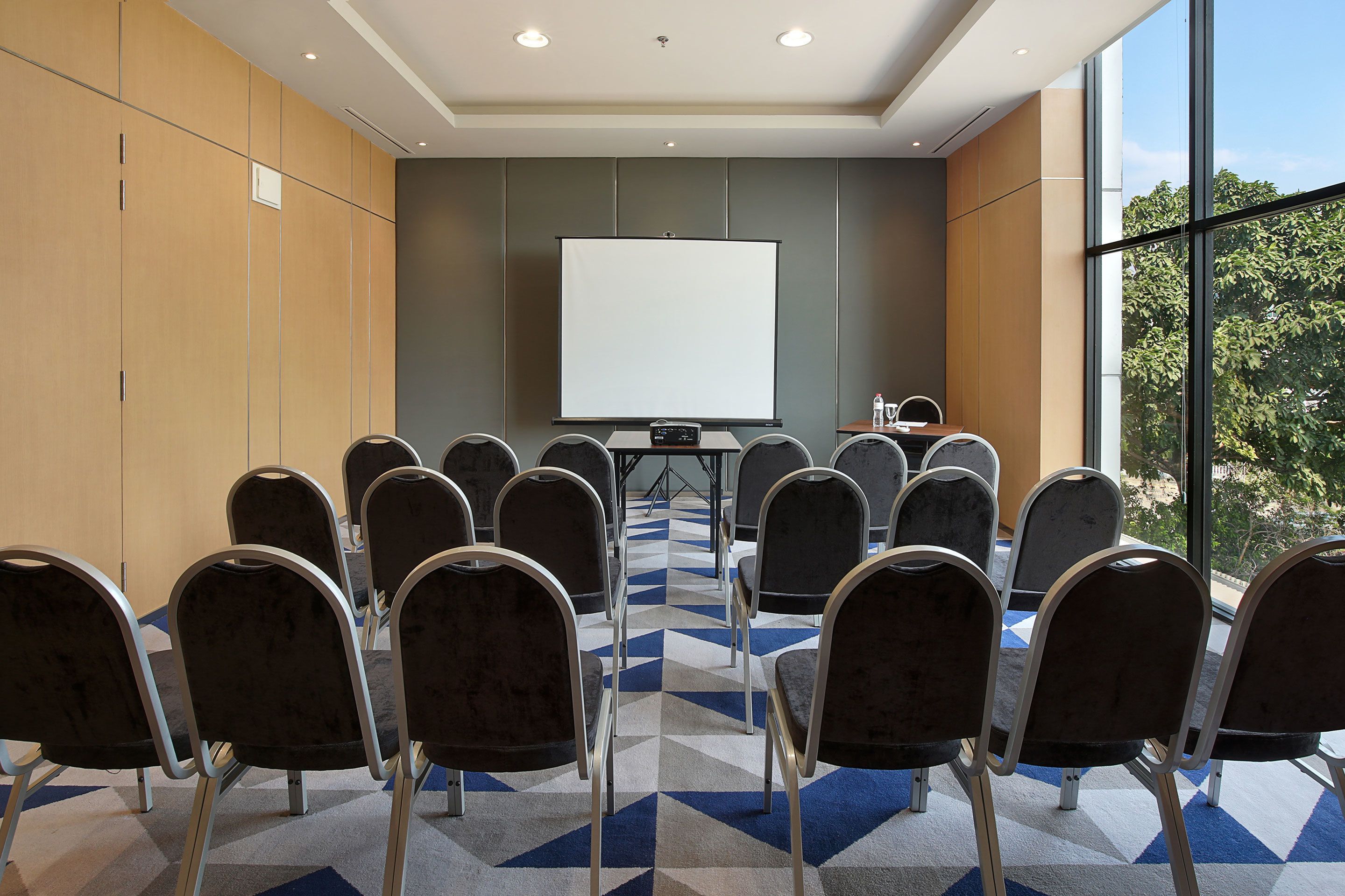 Un salón de conferencias con una pantalla grande, varias sillas y una alfombra estampada.
