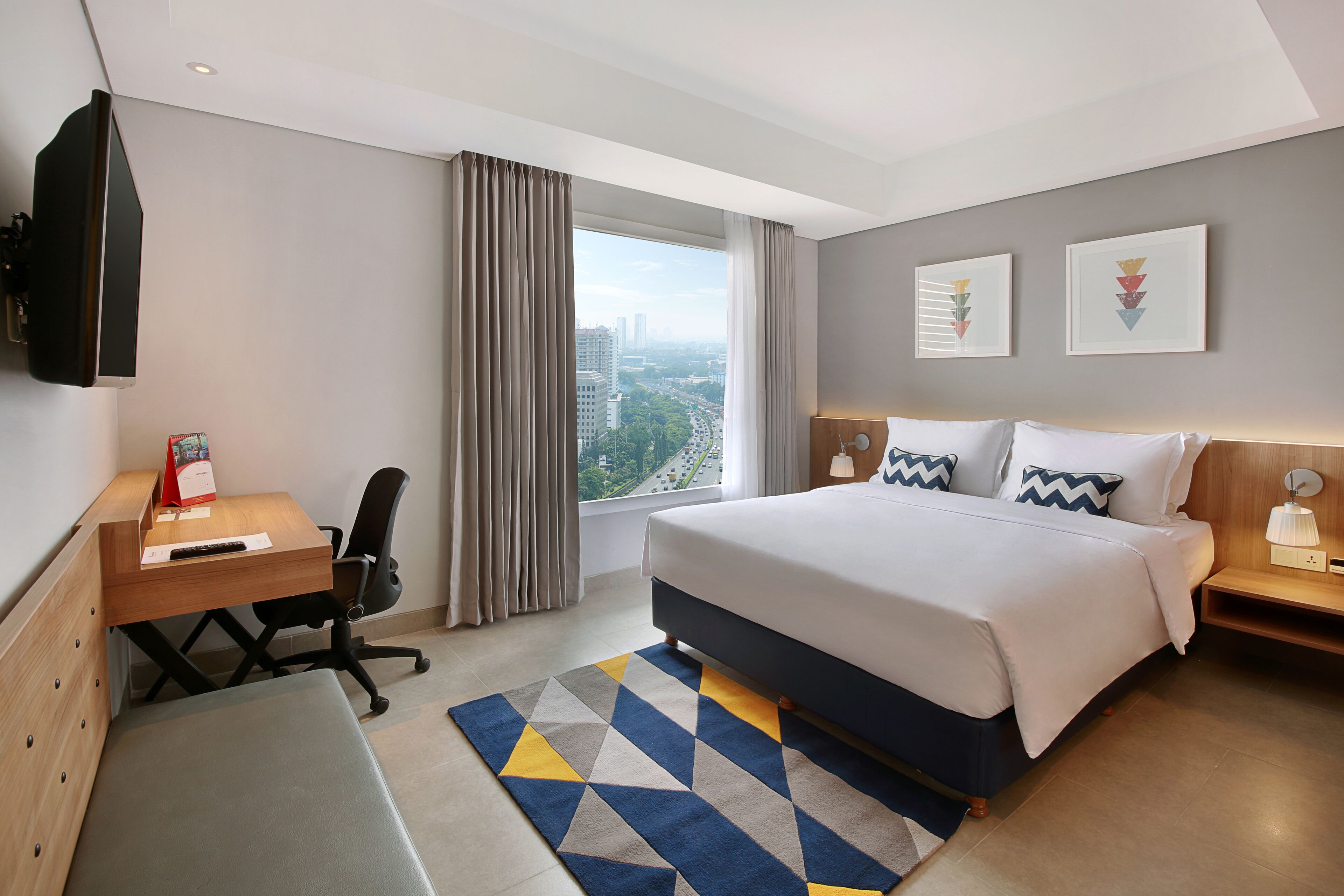 A modern hotel room with a large bed, a desk, a chair, and a view of the city.
