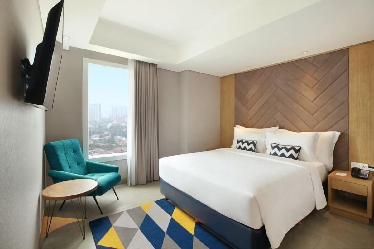 A hotel room with a large bed, a television mounted on the wall, a window with curtains, a teal armchair, and a small table.