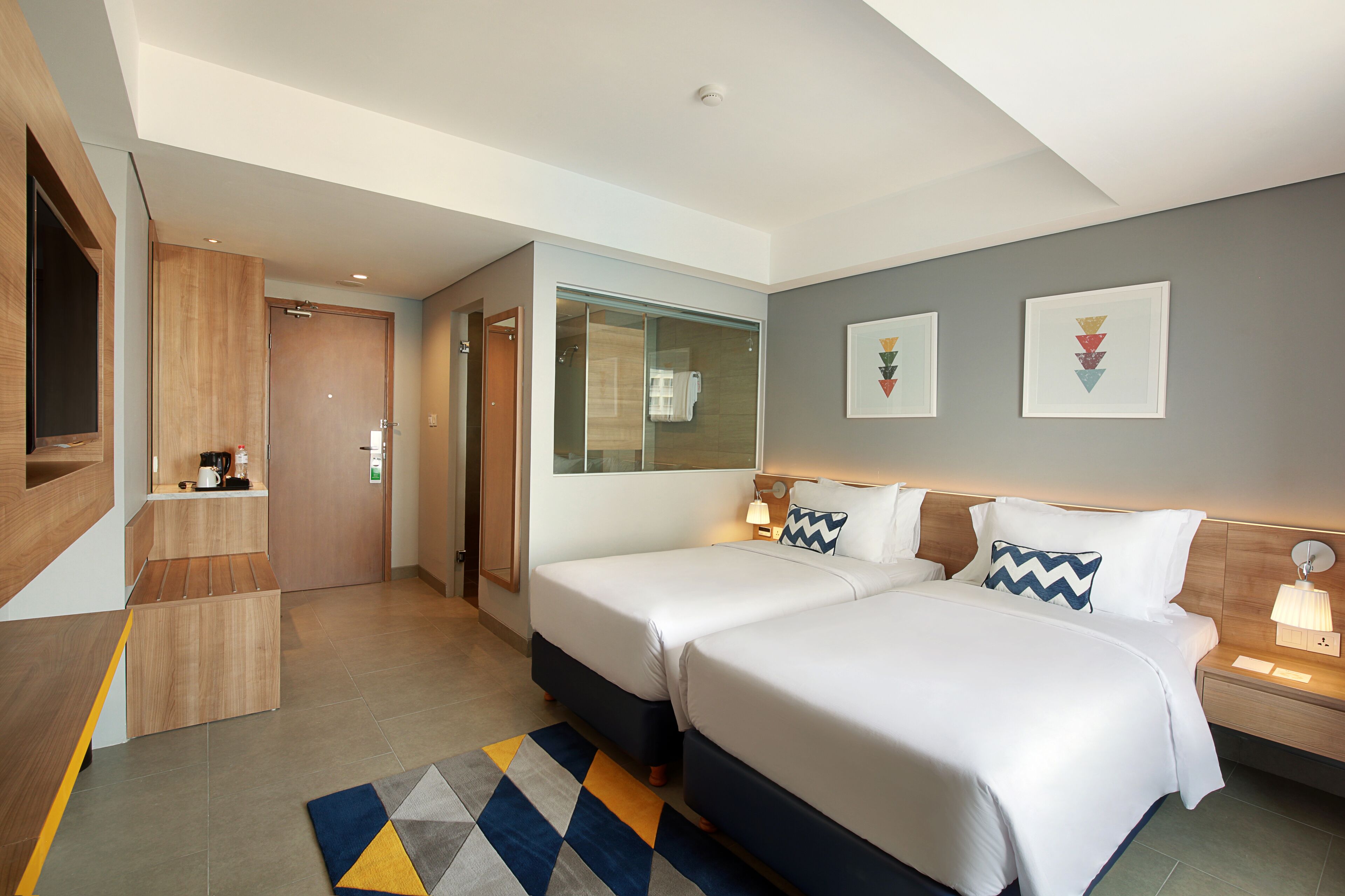 A modern hotel room with two beds, a wooden wardrobe, a small kitchenette, and a large window.