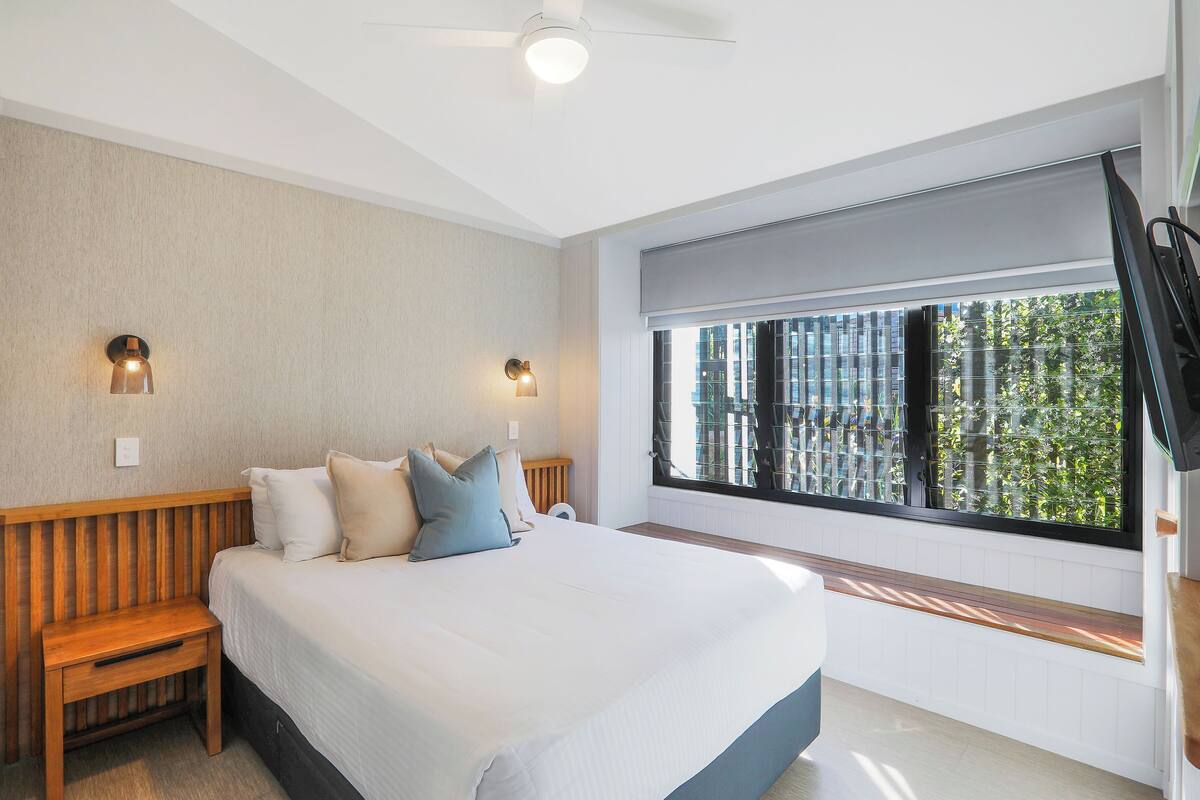 A bedroom with a large bed, two bedside tables, a ceiling fan, and a window with a view of buildings and trees.