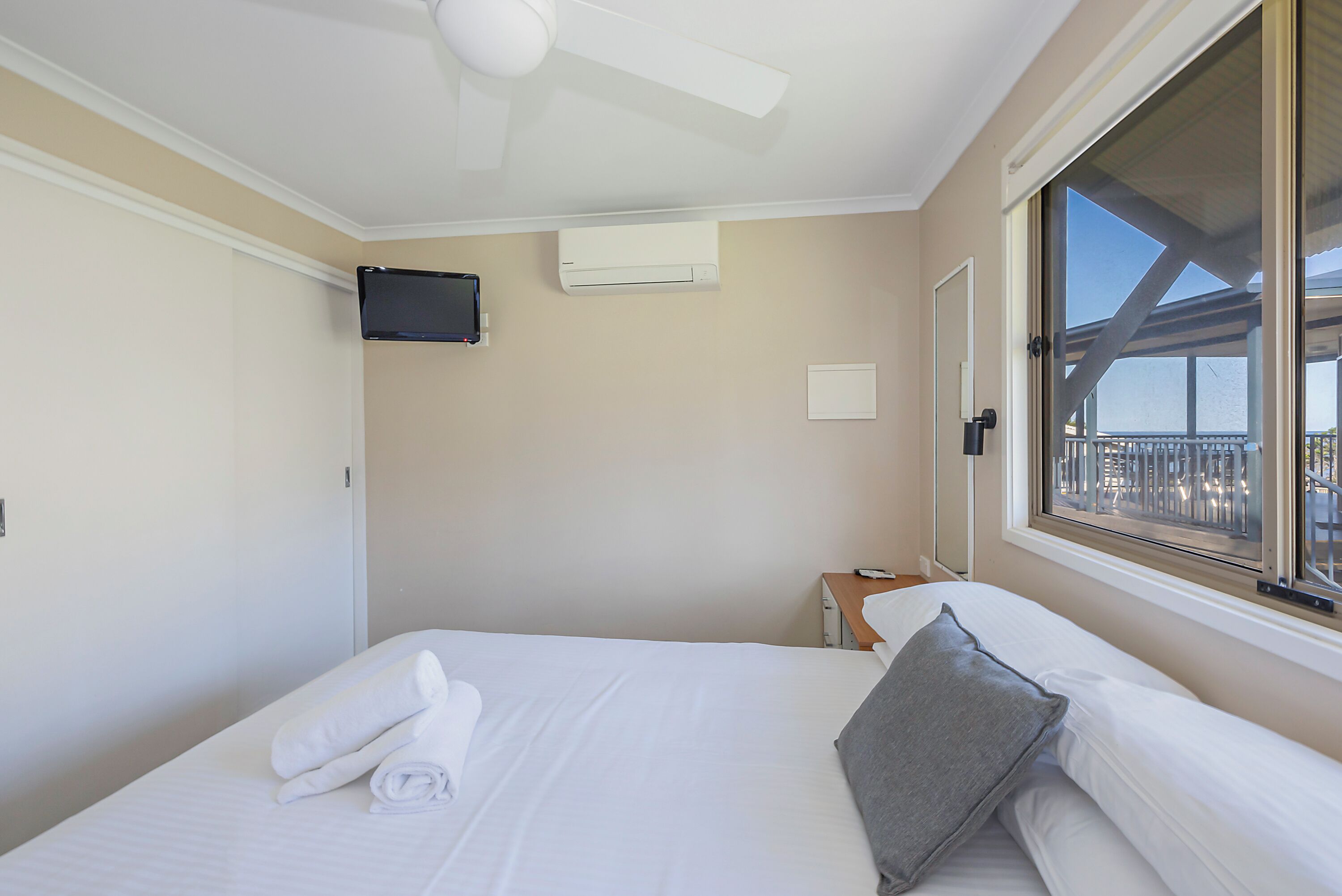 A hotel room with a bed, a flat-screen TV mounted on the wall, an air conditioning unit, a window with a view of a balcony, and a ceiling fan.