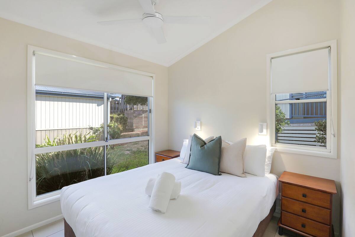 A bedroom with a large bed, two bedside tables, a ceiling fan, and a window with a view of the outside.