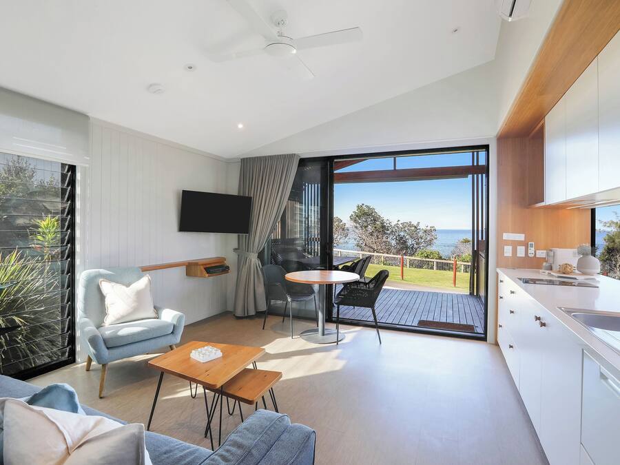 A modern living room with a sofa, armchair, and a small table. There is a balcony with a view of the sea.