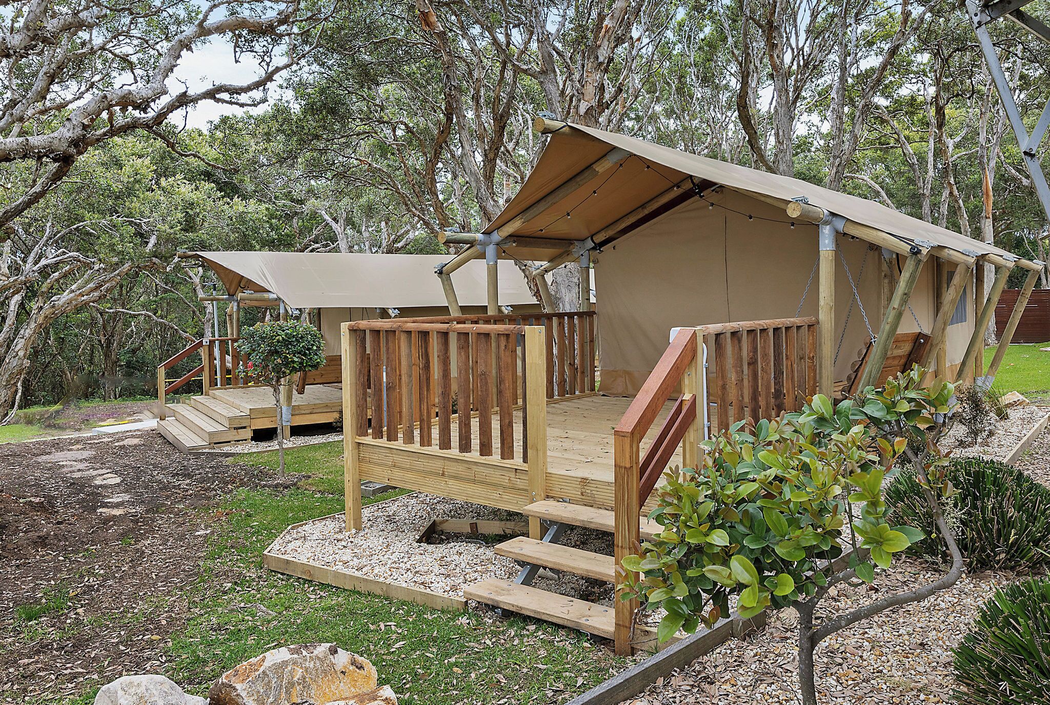 A wooden cabin with a deck and a covered porch, surrounded by trees and gravel pathways.