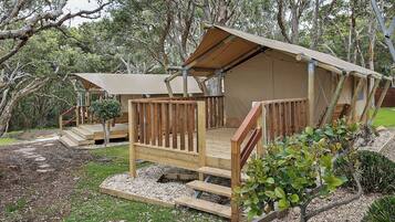 A wooden cabin with a deck and a covered porch, surrounded by trees and gravel pathways.