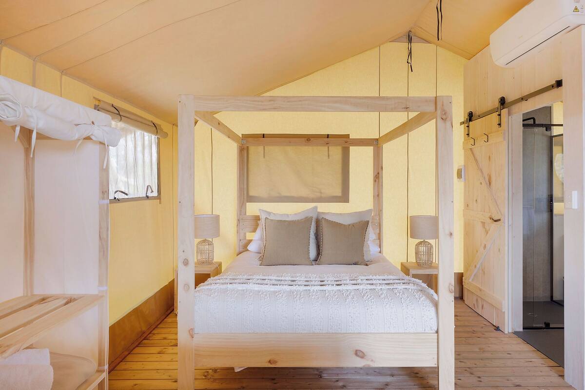 A wooden four-poster bed with a white bedspread and two pillows, a small bedside table with a lamp, and a wooden floor.