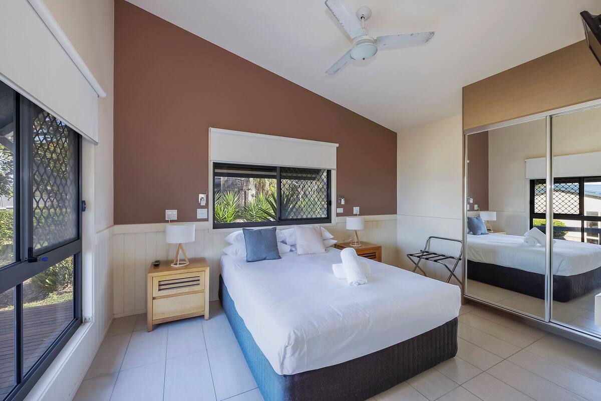 A modern bedroom with a large bed, a nightstand, a lamp, a ceiling fan, and a large mirror.
