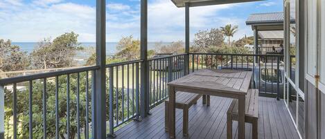 A wooden deck with a table and bench, overlooking a coastal area with trees and a building in the background.