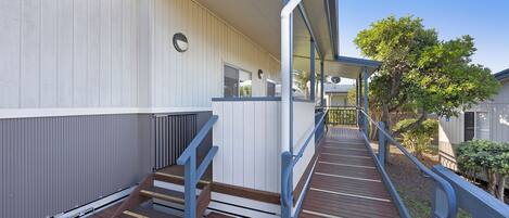 A wooden deck with a metal railing leading to a building with a white exterior and a blue roof.