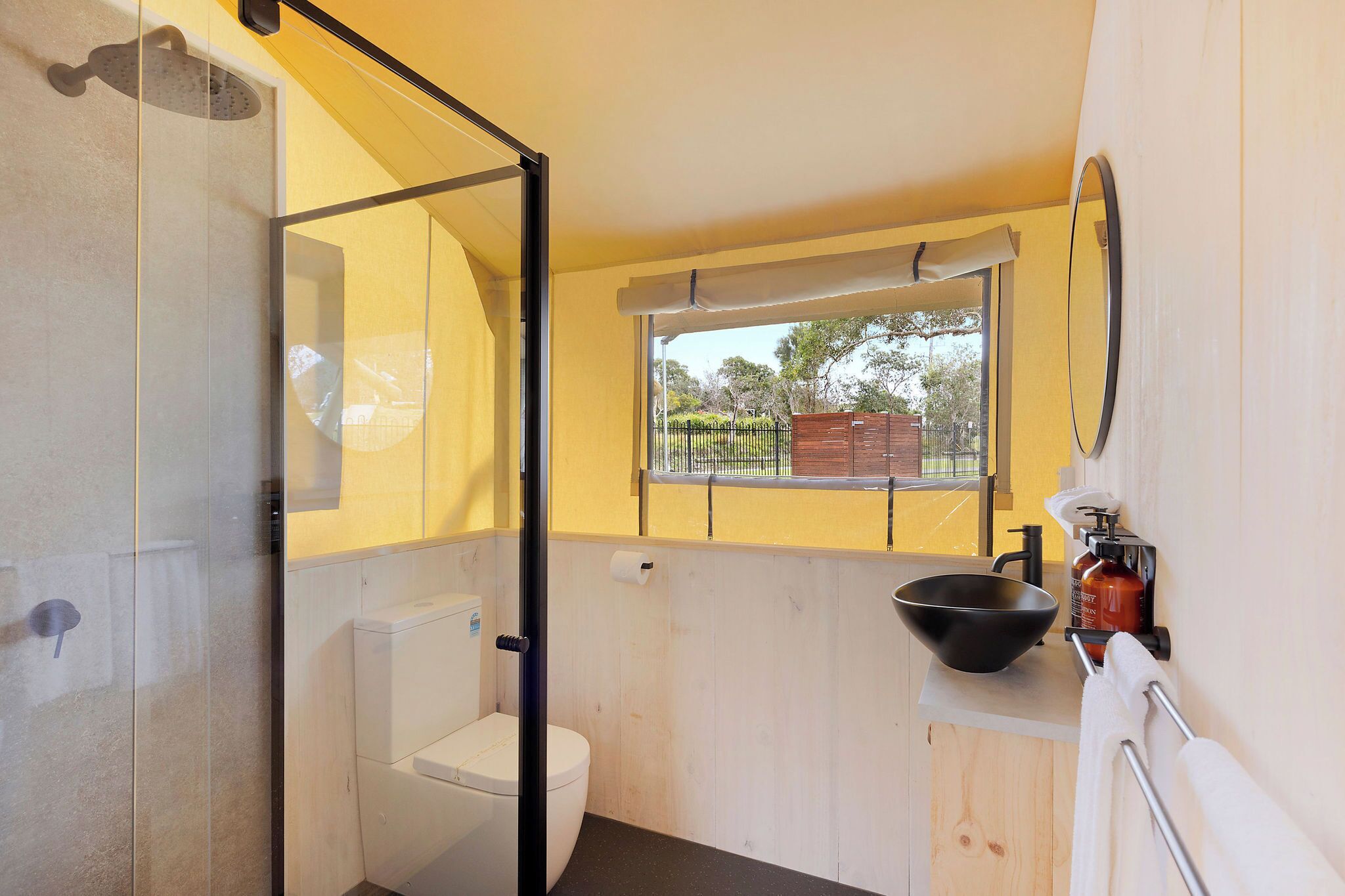 A compact bathroom with a glass shower enclosure, a toilet, a sink with a basin, and a window providing a view of the outside.