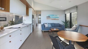 A modern kitchen with white cabinets, a wooden countertop, and a built-in oven. There is a dining area with a round wooden table and black wicker chairs. A blue sofa and a chair are in the living area, with a painting of a beach scene on the wall. - Reflections Bonny Hills - Holiday Park (Bonny Hills)