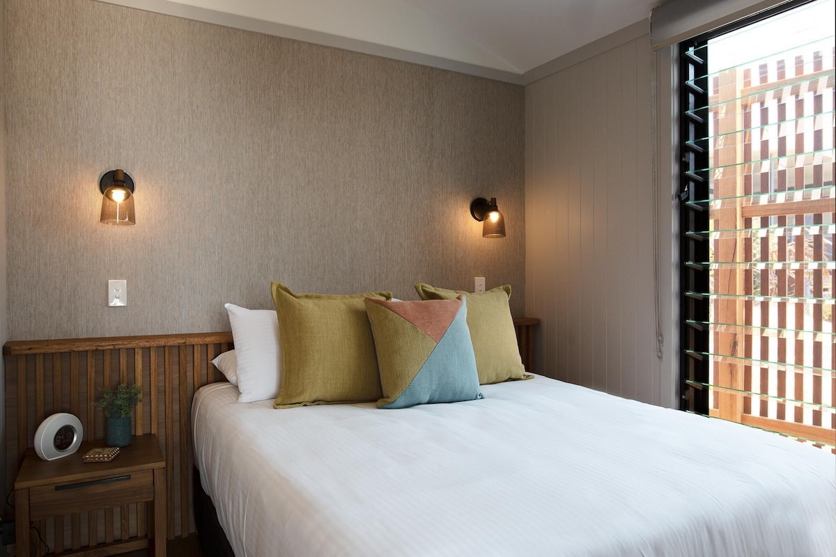A neatly made bed with pillows, two wall-mounted lights, a nightstand with a plant, and a window with a wooden lattice.