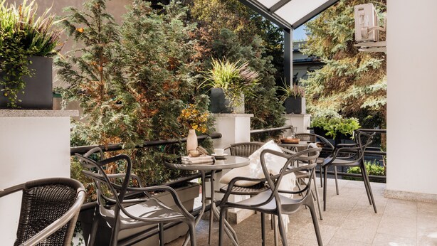Une terrasse extérieure avec des chaises et des tables en osier, entourée de plantes en pot et offrant une vue sur un bâtiment.